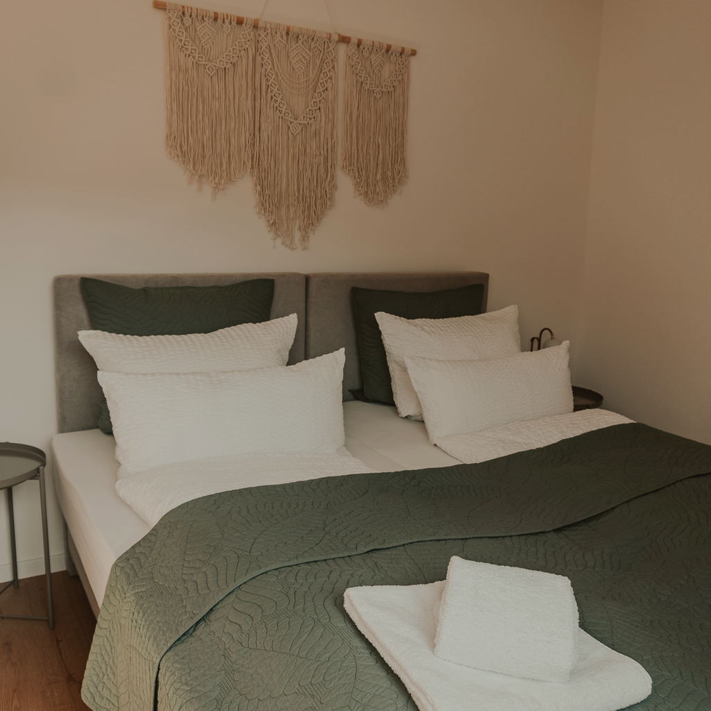 Doppelbett mit dunkelgrüner Tagesdecke, weißen Kissen und Boho-Deko